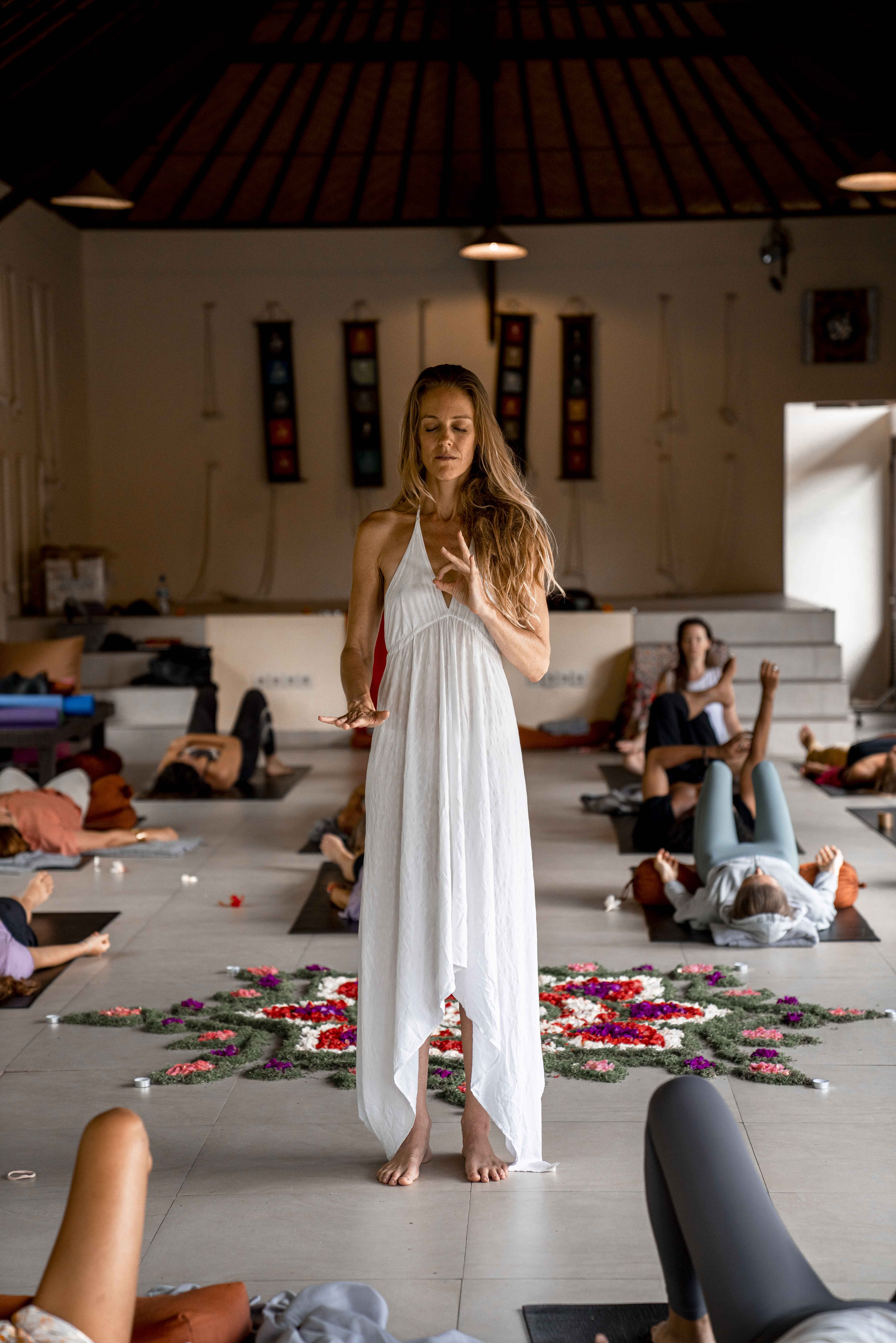 Marika Daya leading a group Kundalini transmission session, standing barefoot in white before a flower mandala while participants rest on mats around her